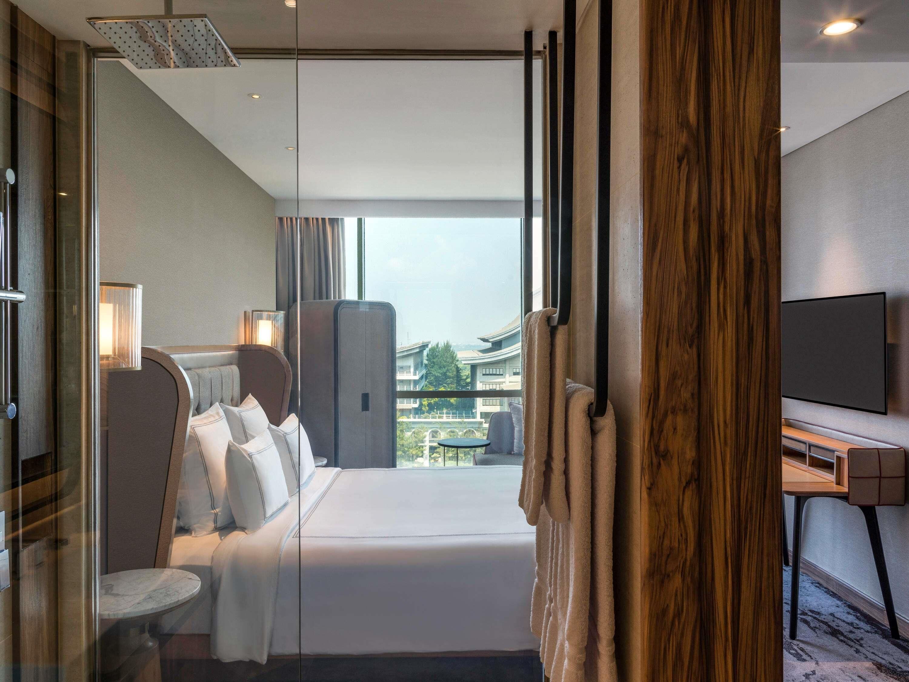 A modern hotel room with a large bed, a TV, and a view of the cityscape.