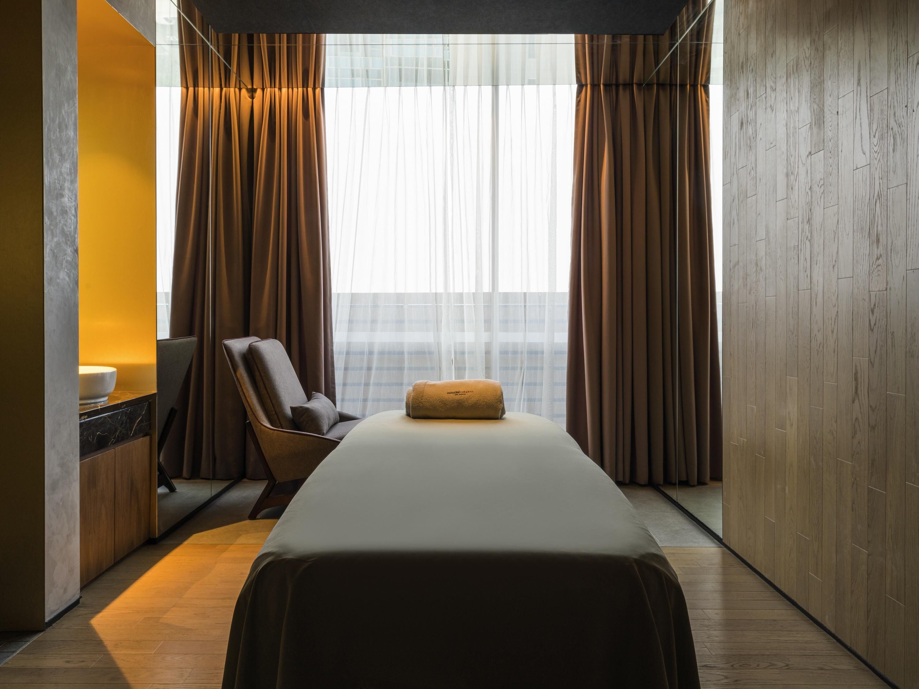 A modern hotel room with a large bed, a seating area with a chair and a small table, and a view of the city through sheer curtains.