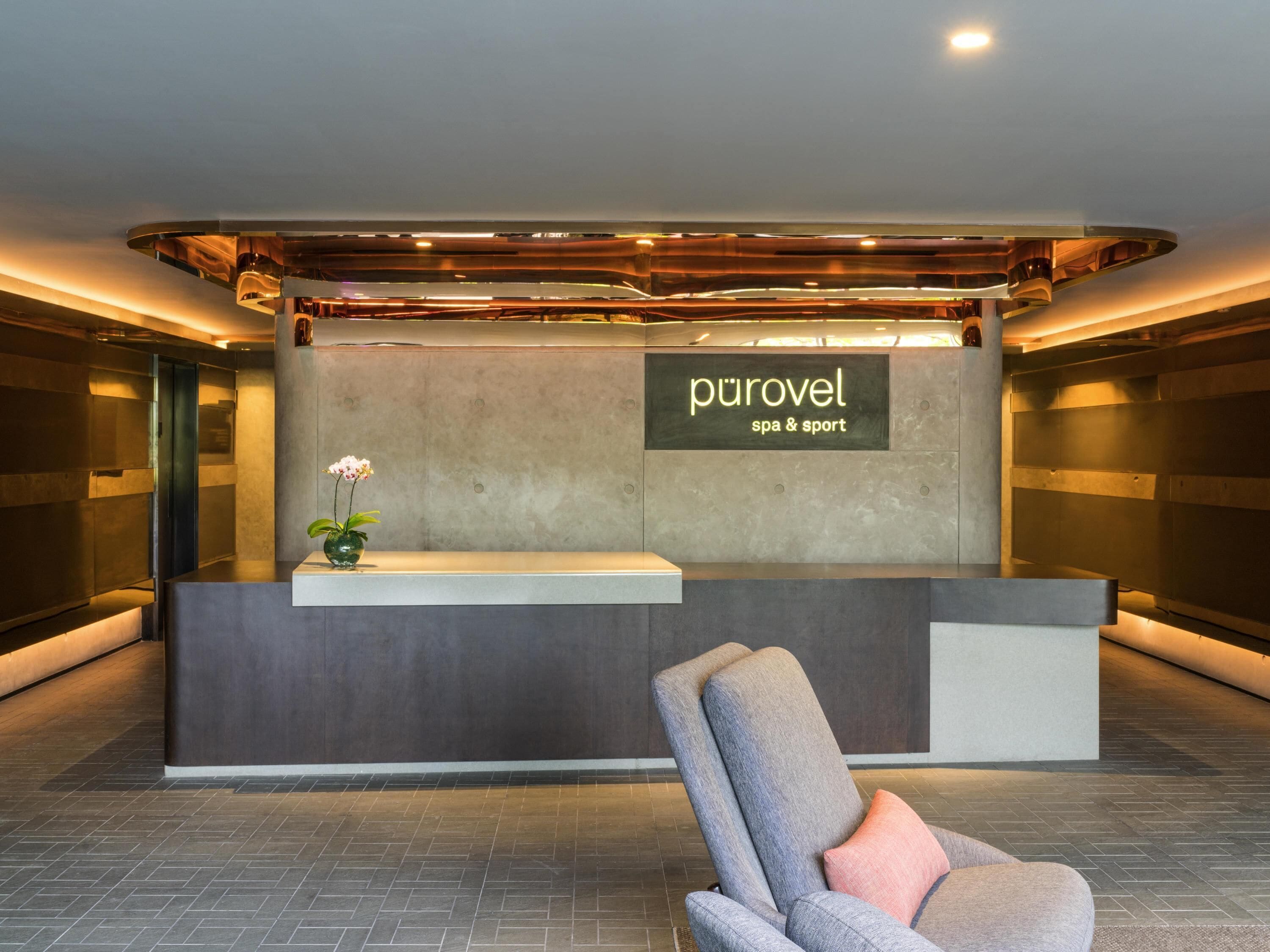 Un mostrador de recepción con un cartel que dice 'purovel spa & sport' en un vestíbulo moderno con una silla y un jarrón de flores.