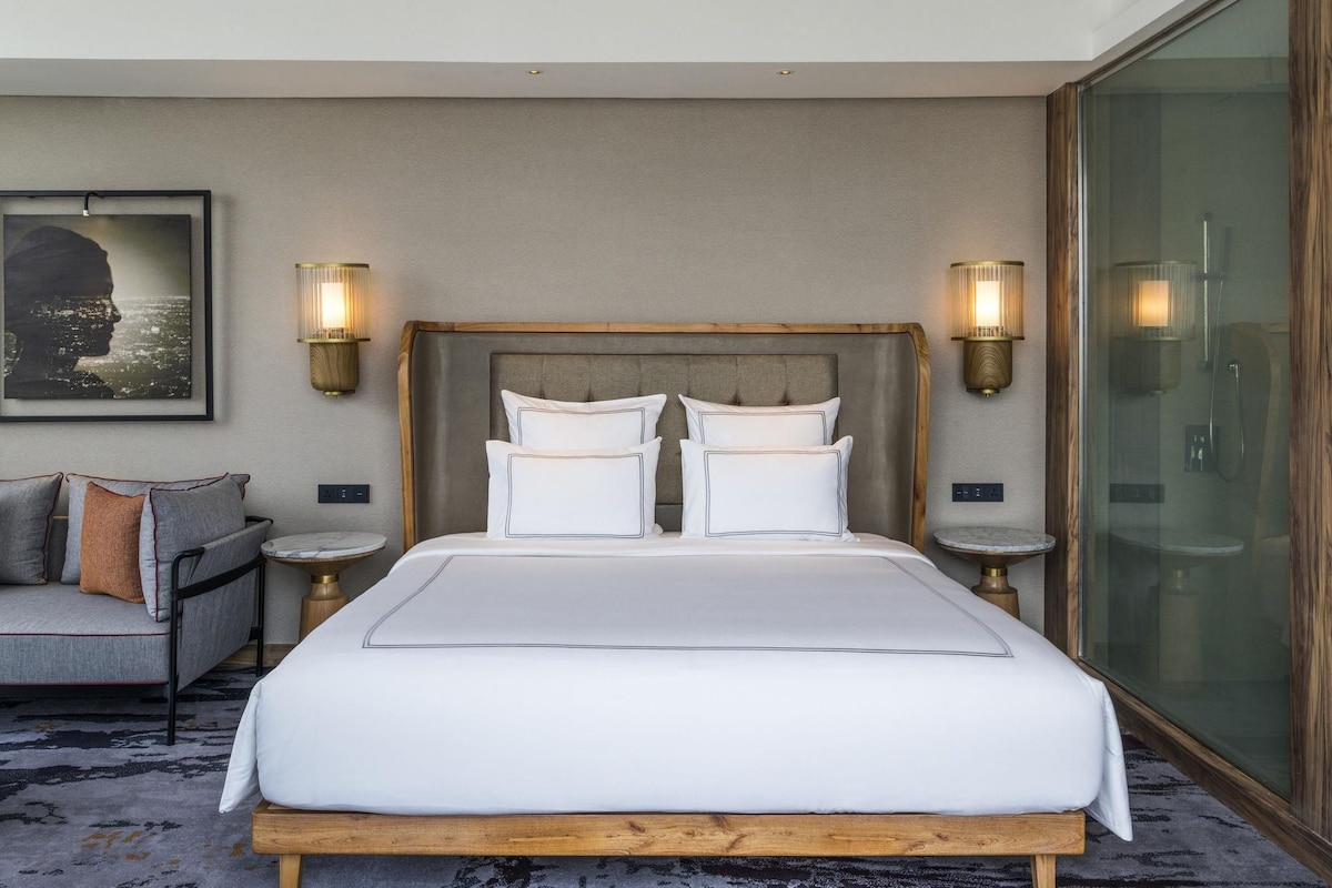 A hotel room with a large bed, two bedside tables, a sofa, a framed picture on the wall, and a large window.