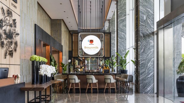 Un moderno vestíbulo de hotel con mostrador de recepción, zona de estar y un letrero para Brownmilk 24 Hour Deli.