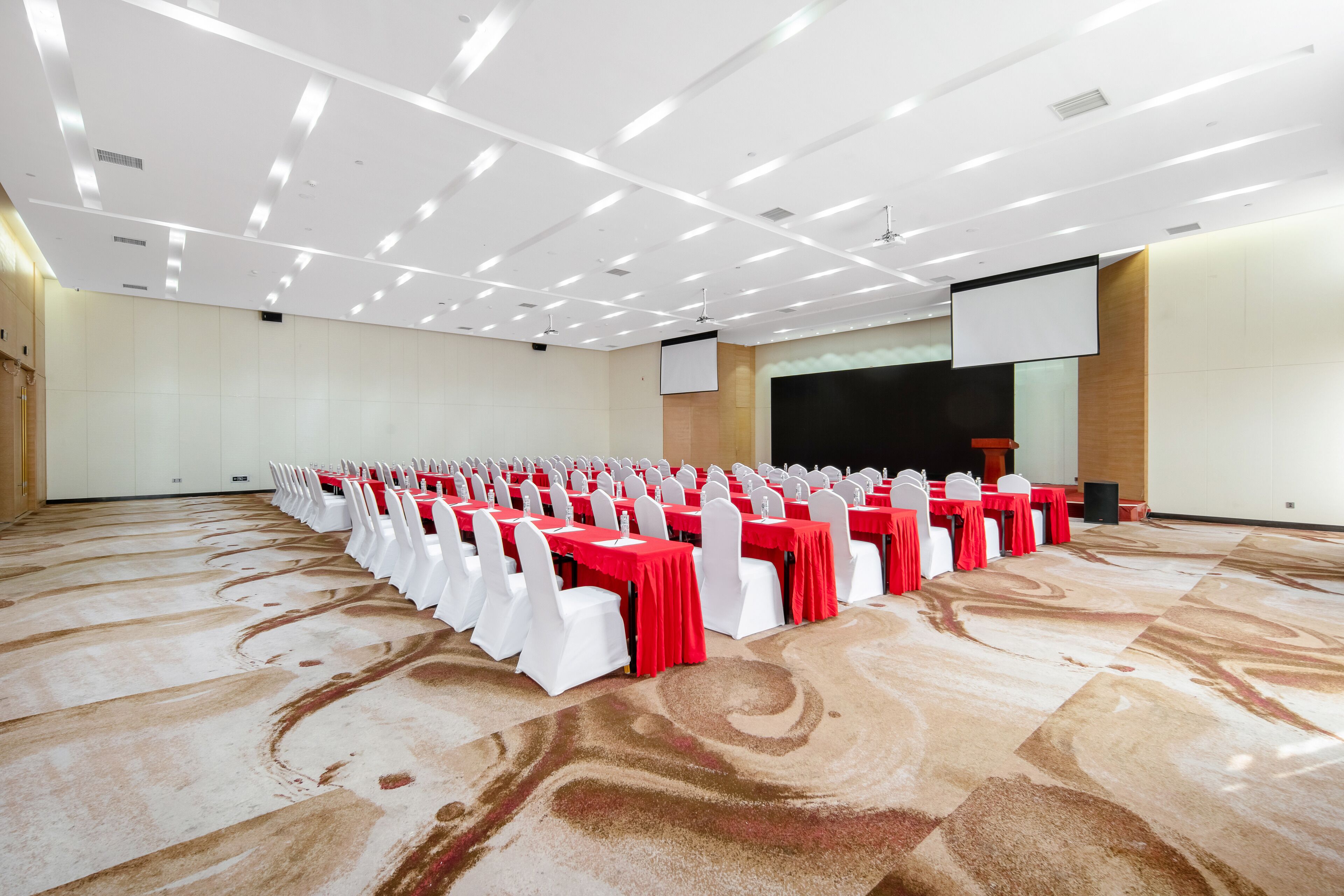 Un amplio salón de conferencias con filas de mesas y sillas, una pantalla de proyector y un atril.