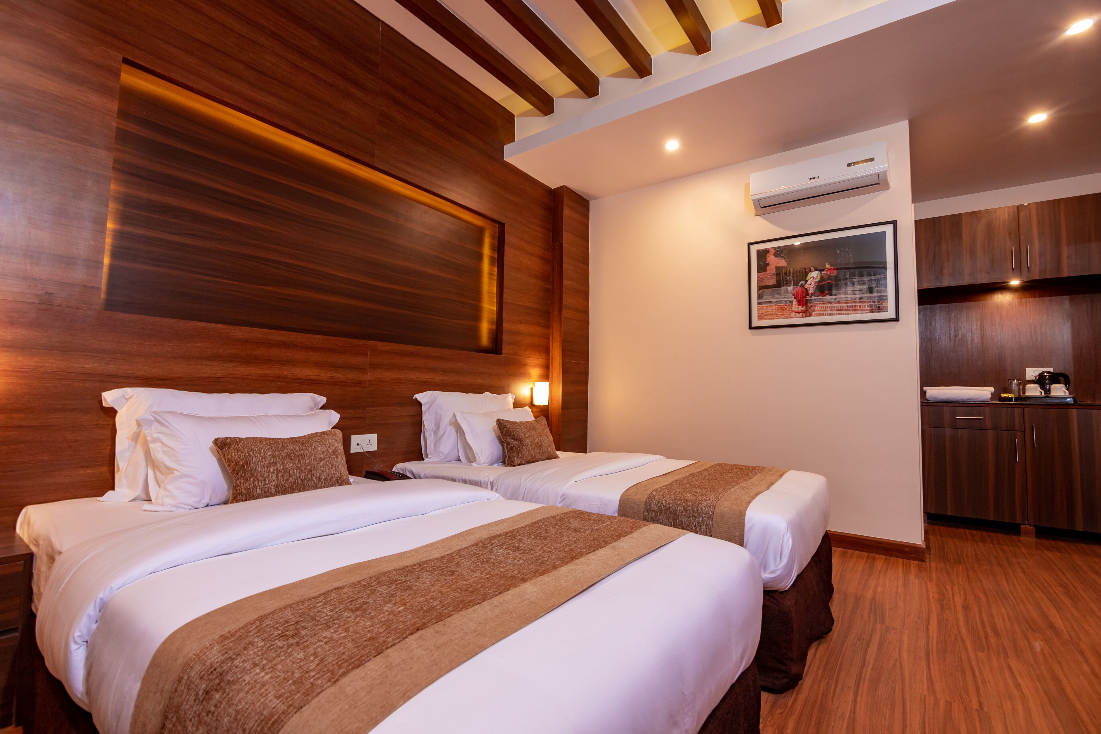 Habitación de hotel con dos camas, paneles de madera, aire acondicionado montado en la pared, un cuadro enmarcado y una zona de kitchenette.