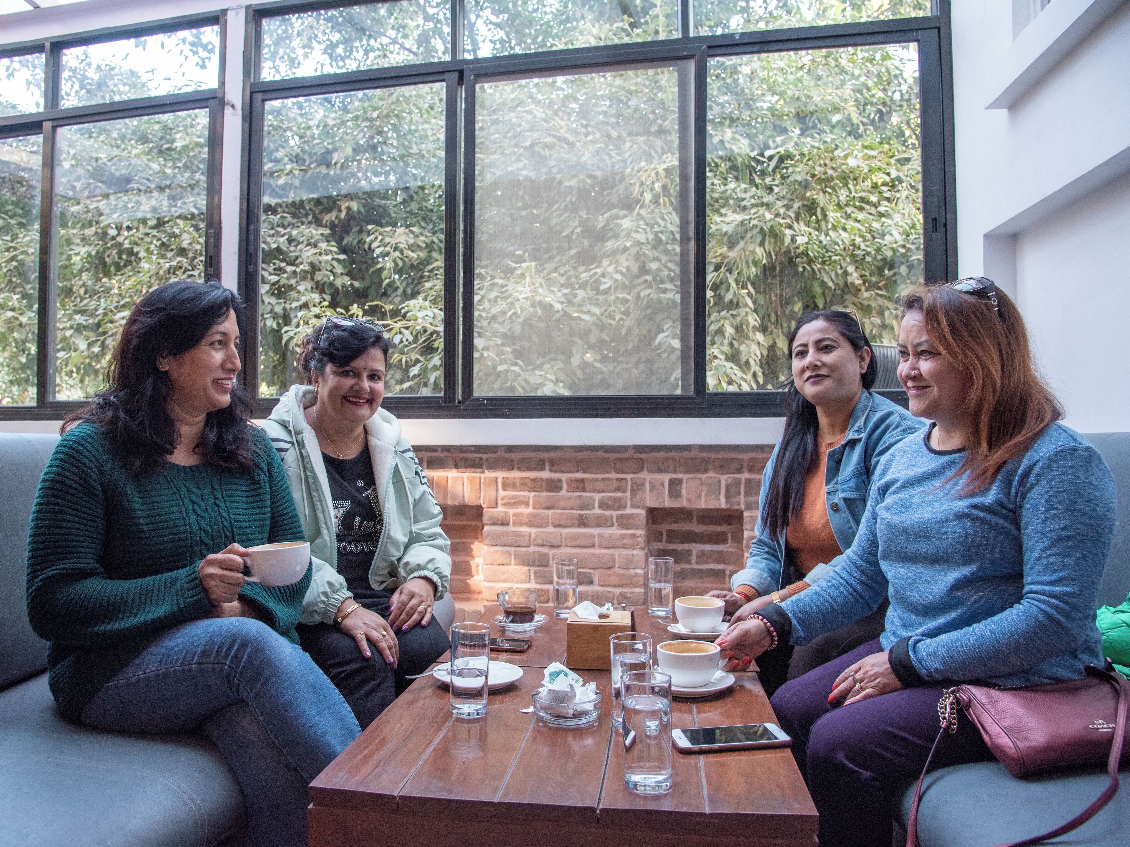 A group of people sitting around a wooden table with drinks and a smartphone, in a room with large windows and a brick wall.