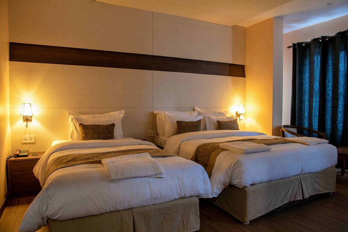 A hotel room with two beds, each with a pillow, a bedside table with a lamp, and a chair.