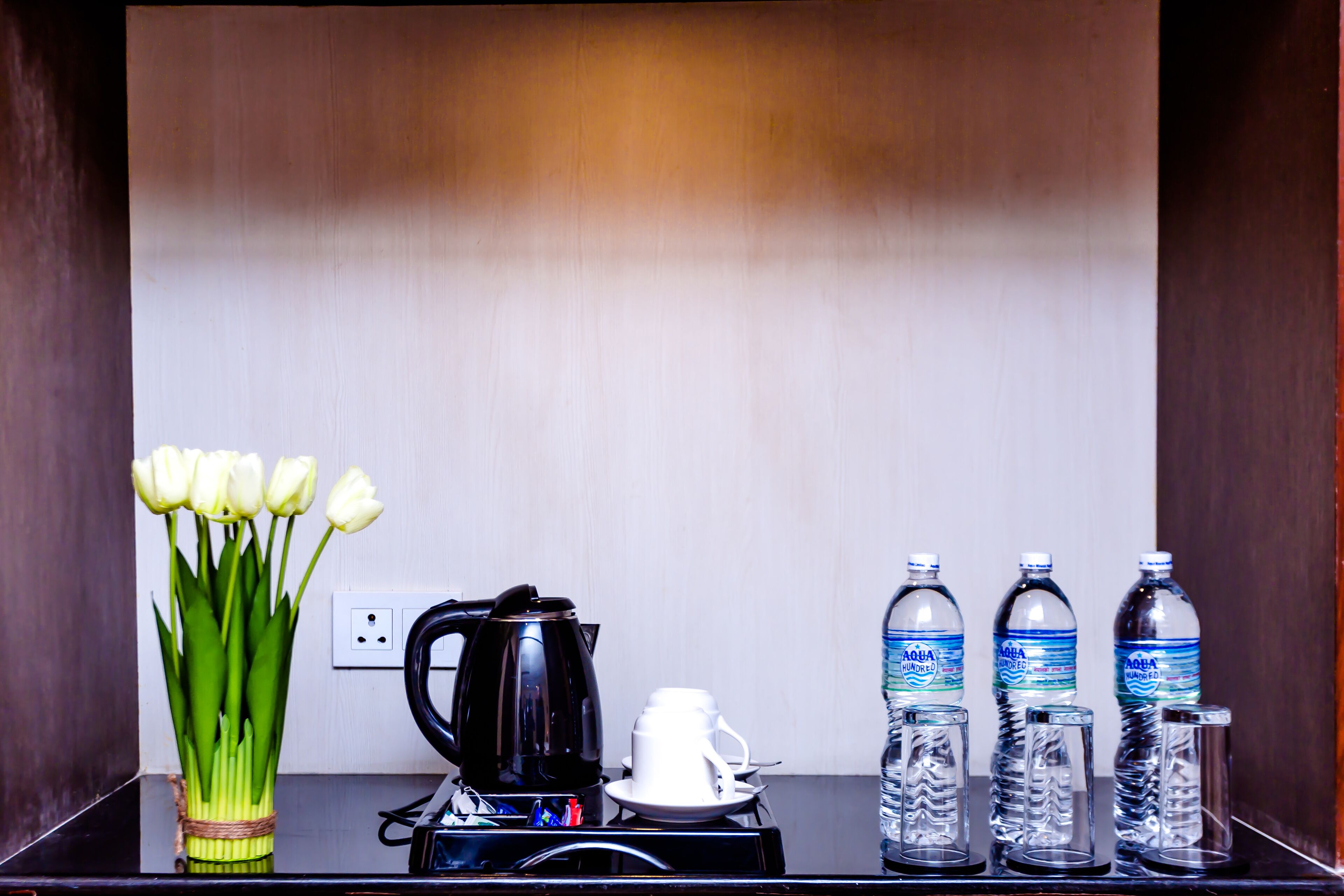 Habitación de hotel con una mesa negra, un jarrón de tulipanes blancos, una cafetera negra, tazas blancas y tres botellas de agua.