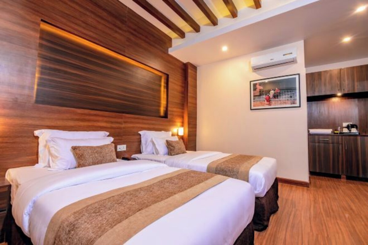 Habitación de hotel con dos camas, cabeceros de madera, un aire acondicionado de pared, un cuadro enmarco y una kitchenette con armarios oscuros.