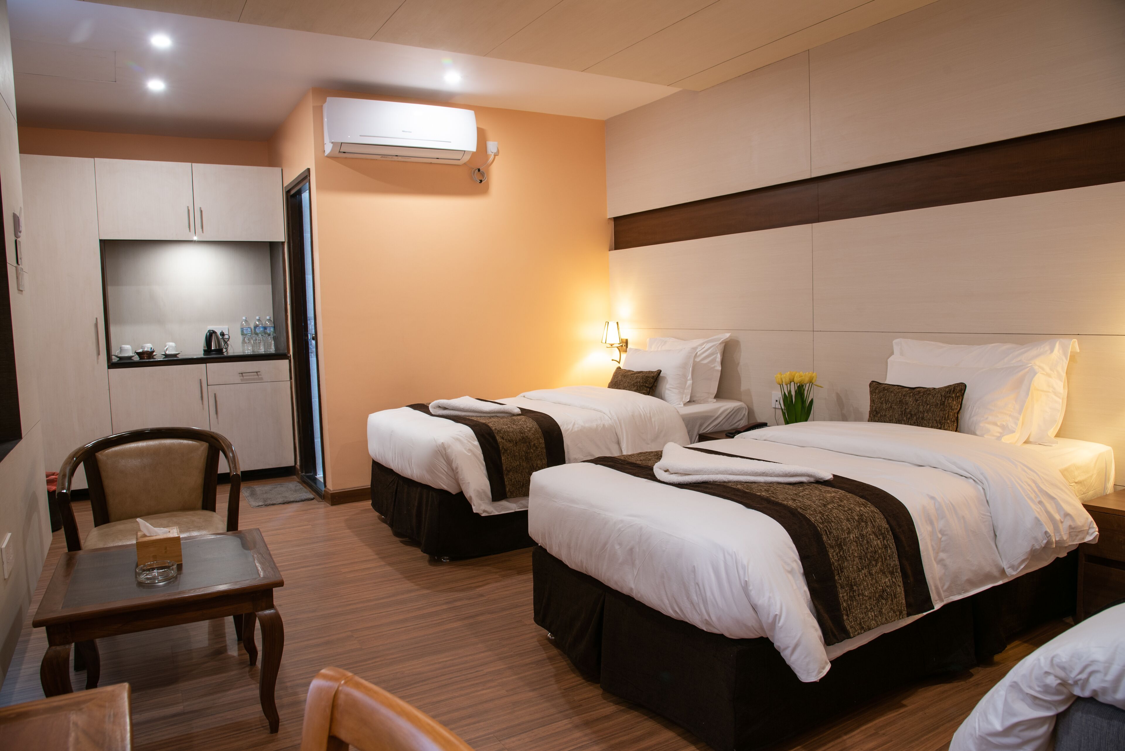 Habitación de hotel con dos camas, una mesita, una silla, kitchenette y aire acondicionado.
