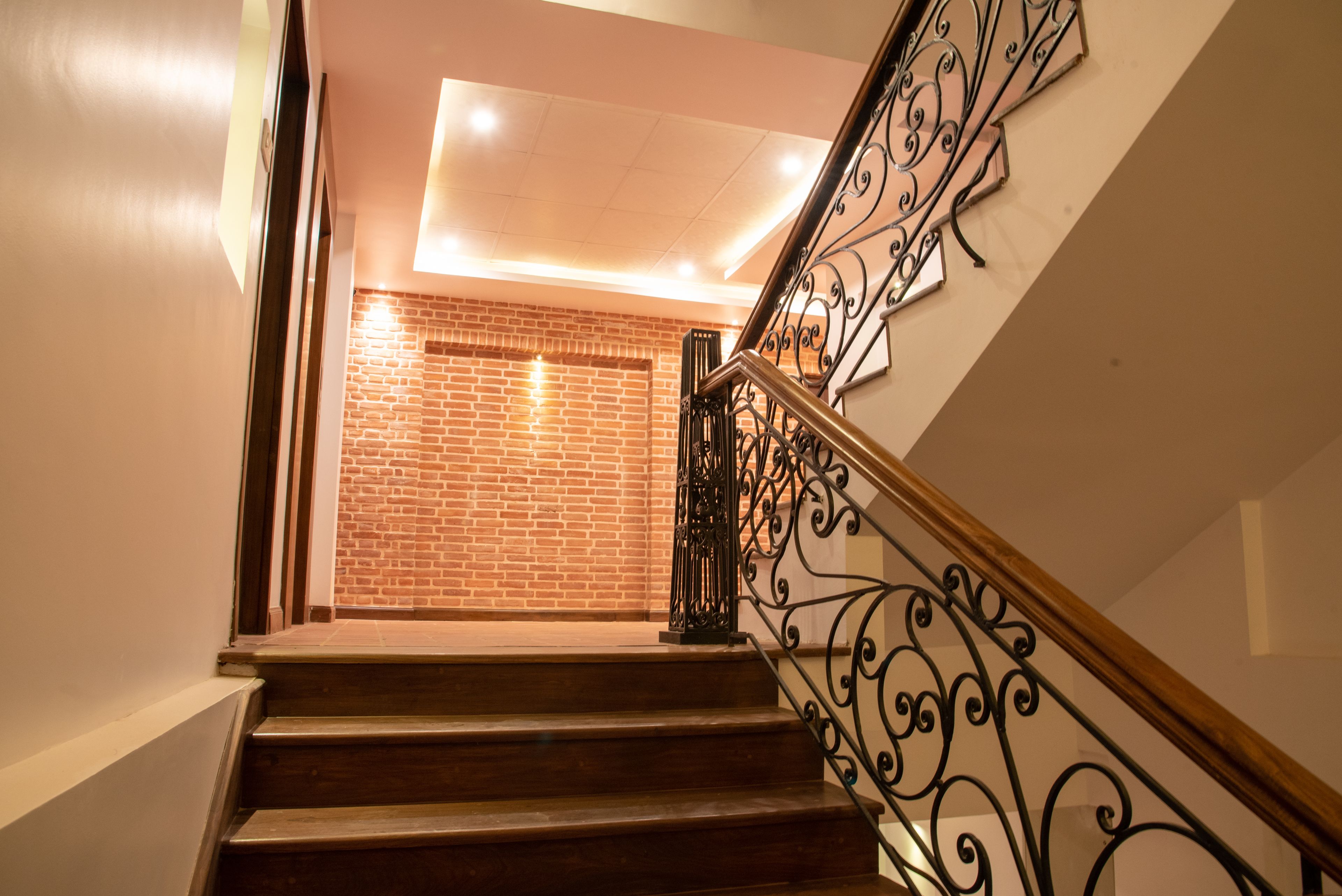 A staircase with wooden steps and black wrought iron railings, leading to a room with a brick wall and recessed ceiling lighting.