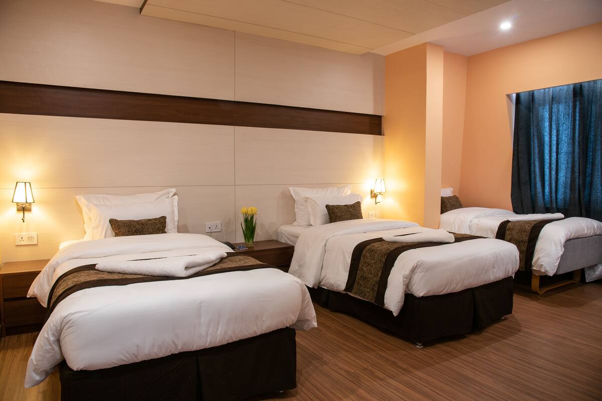 A hotel room with three beds arranged in a row, each with a bedside table and lamp.