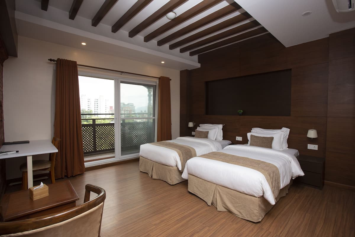 A hotel room with two beds, a desk, and a chair. There is a balcony with curtains and a view of buildings.