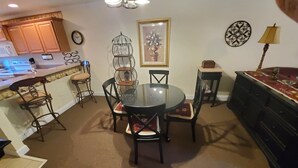 A dining area with a round table and chairs, a kitchen with wooden cabinets, a clock, and decorative wall art. - Little Black Bear Condo (Helen)