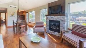 A spacious living room with a stone fireplace, a television mounted above it, and a dining area with wooden furniture. - Two Bedroom Oceanfront Villa - Mayne Island Resort (Mayne Island)