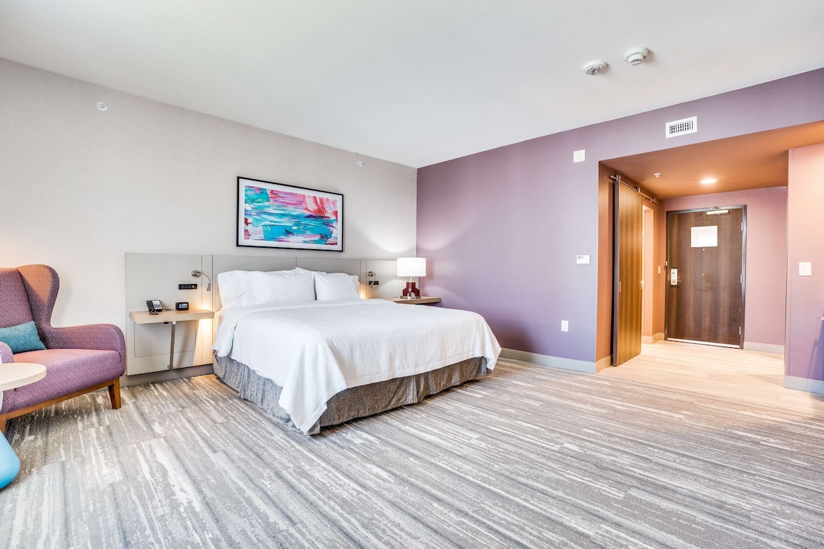 Hotelkamer met een bed, een stoel, nachtkastje, lamp en een schilderij aan de muur.