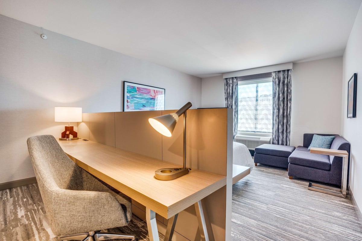 Een hotelkamer met een houten bureau, een stoel, een bank, een lamp en een raam met gordijnen.