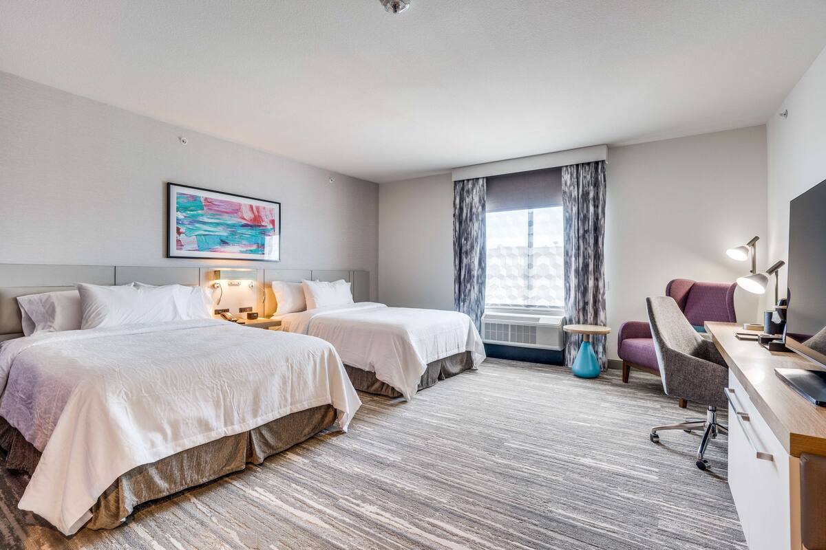 Een hotelkamer met twee bedden, een bureau met stoel, een televisie, een schilderij aan de muur en een raam met gordijnen.