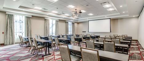 A conference room with multiple tables and chairs, a projector screen, and large windows with curtains.