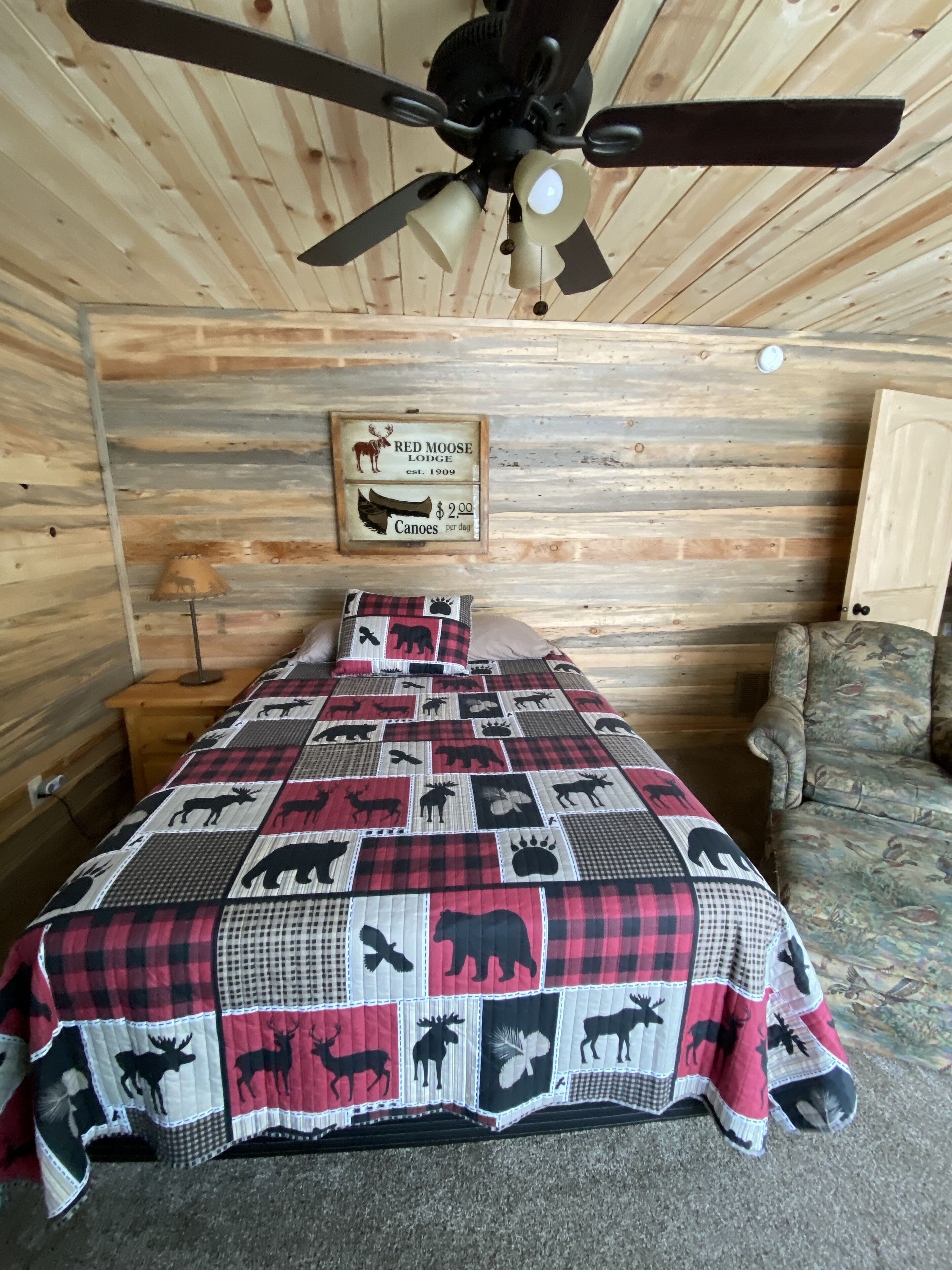 Quarto com cama, ventoinha no teto, parede de madeira, uma placa e um cobertor estampado.