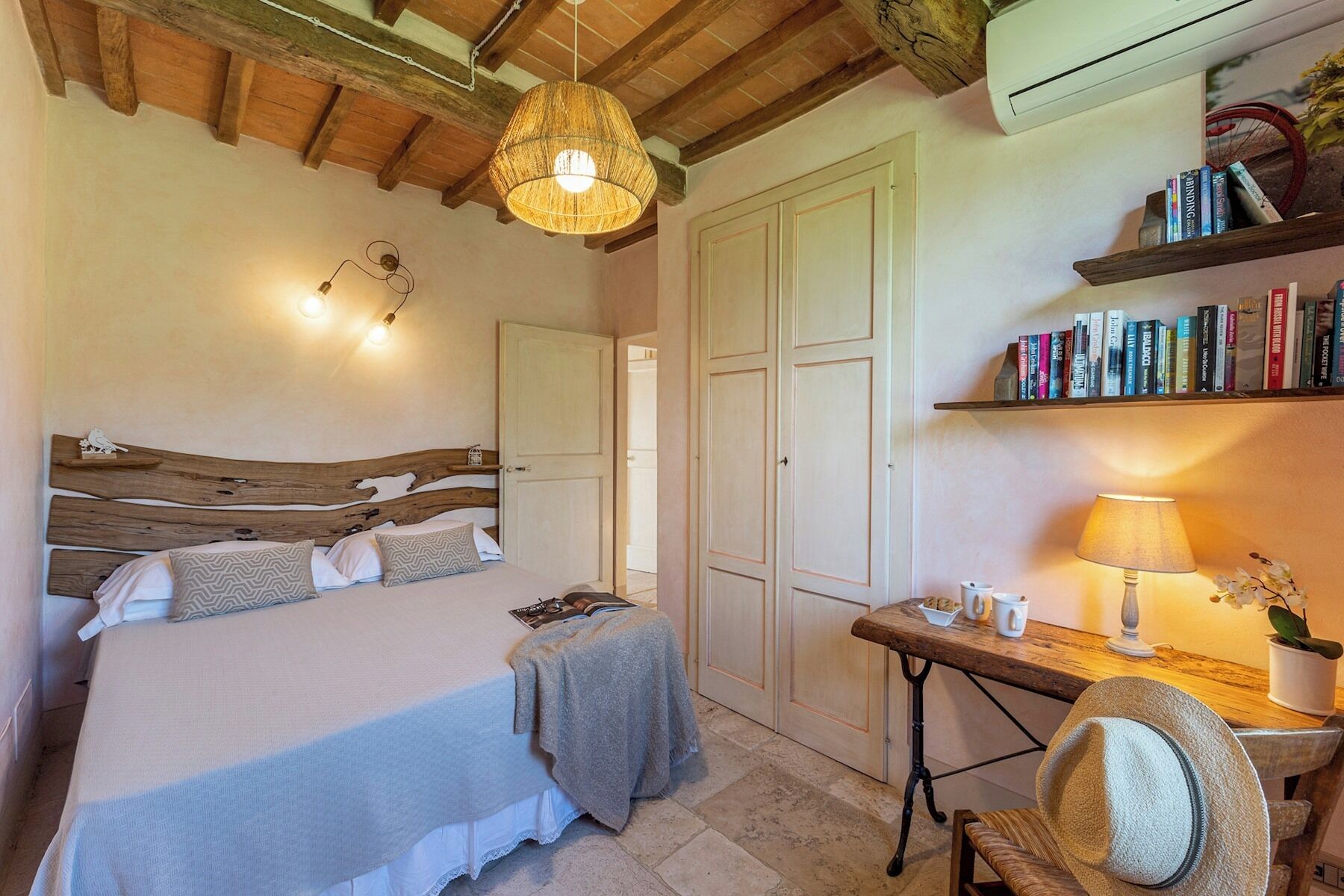 A bedroom with a wooden headboard, a bed with a grey blanket, a bedside table with a lamp, a chair, and a bookshelf with books.