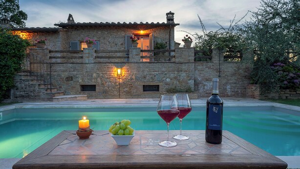 Mesa junto a la piscina con una vela, uvas y copas de vino, frente a un edificio de piedra con luz cálida.