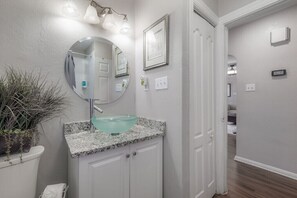 Un baño con lavamanos de vidrio, muebles blancos, un espejo y una planta en maceta.