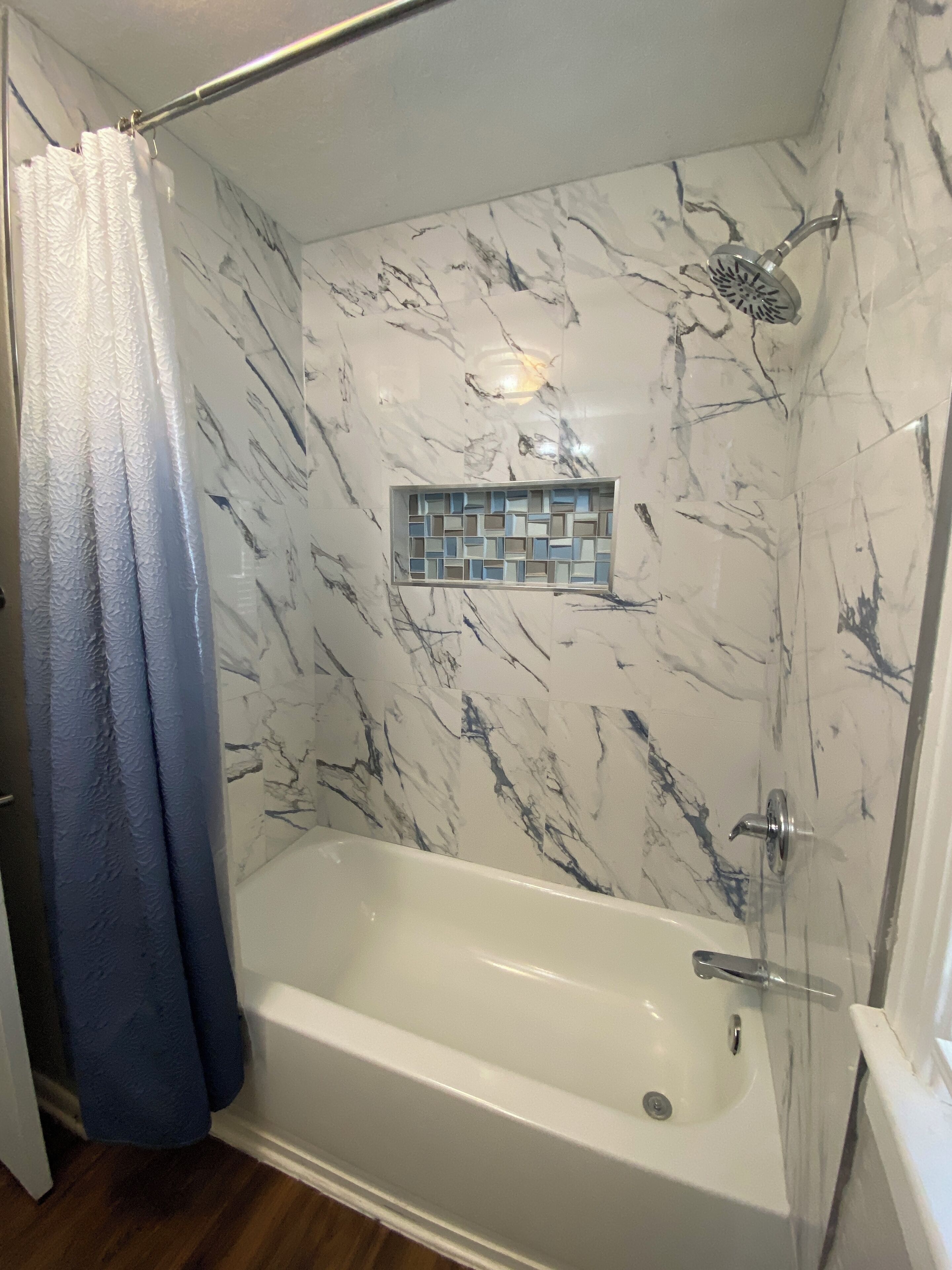 Une douche à l’italienne avec une porte en verre, une baignoire blanche et un mur décoré d’un motif marbré.