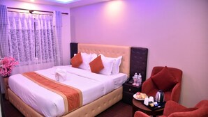 A hotel room with a large bed, a nightstand with a phone and water bottles, a red armchair, and a vase of pink flowers. - Marinha Airport Hotel (Kathmandu)
