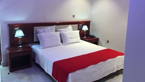 A neatly made bed with white bedding and a red blanket, flanked by two bedside tables with lamps, in a room with white walls and a wooden headboard. - Salerato Boutique Hotel (Gaborone)