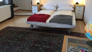 Een net opgemaakt bed met een rood-grijs deken, twee nachtkastjes met lampen, een kamerbreed tapijt met een patroon en een wereldbol op een bureau.