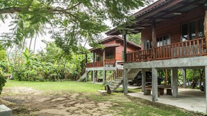 A two-story wooden house with a balcony, surrounded by lush greenery and a paved driveway.