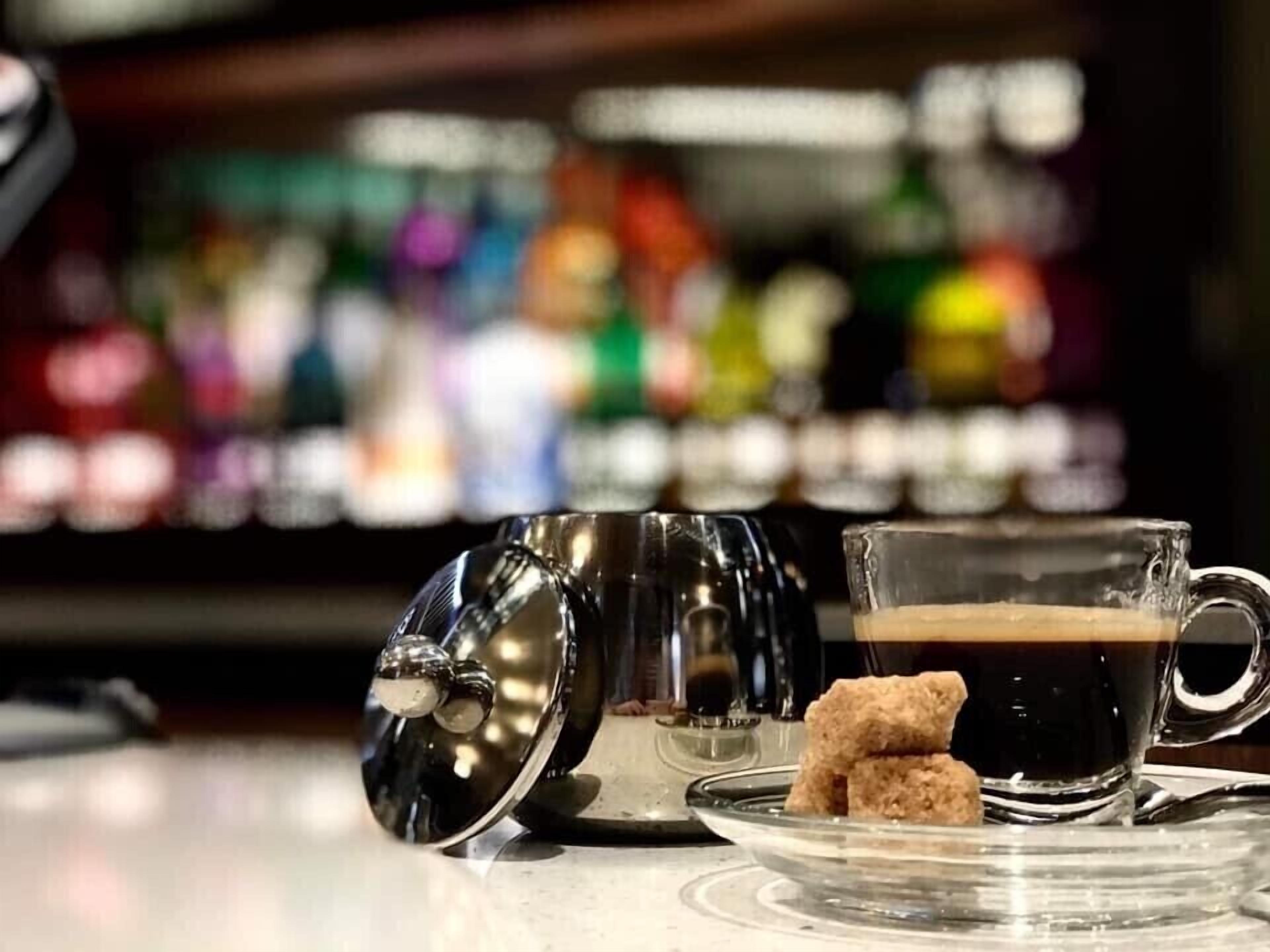 A bar setting with a coffee cup, a glass of dark liquid, and a sugar packet on a clear glass plate.