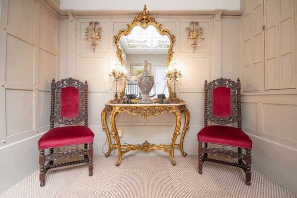 A room with two ornate chairs, a gold-framed mirror, and a table with a red cloth.