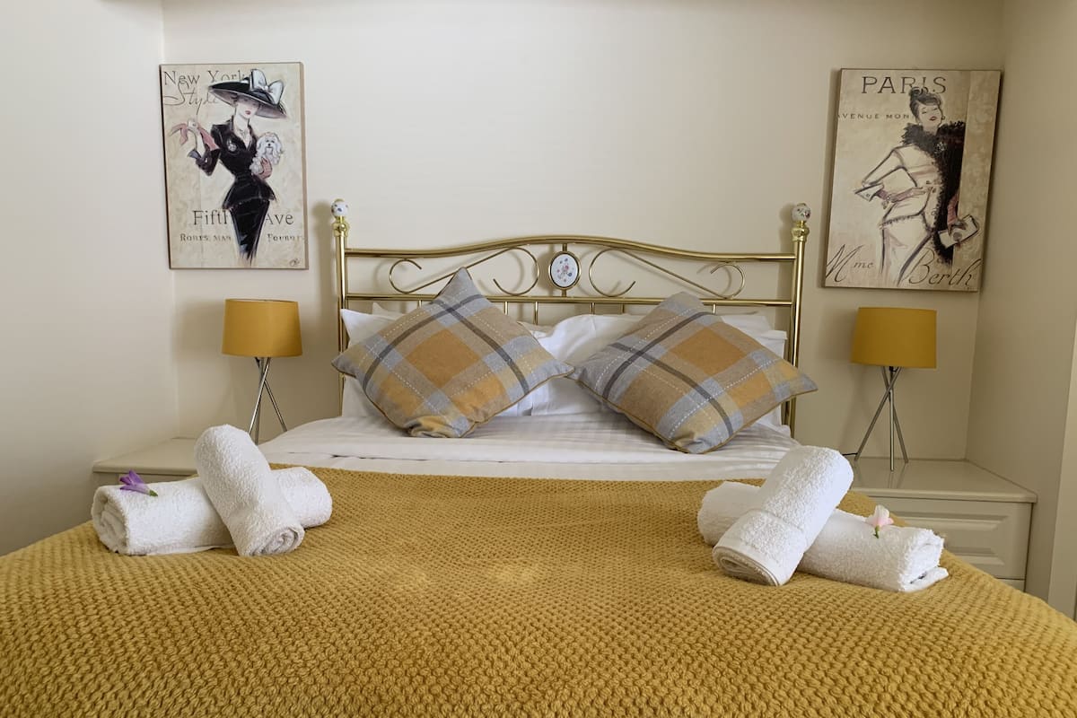 A neatly made bed with a yellow knitted blanket, two plaid pillows, and two white towels with purple flowers. There are two wall-mounted lamps and two framed pictures on the walls.