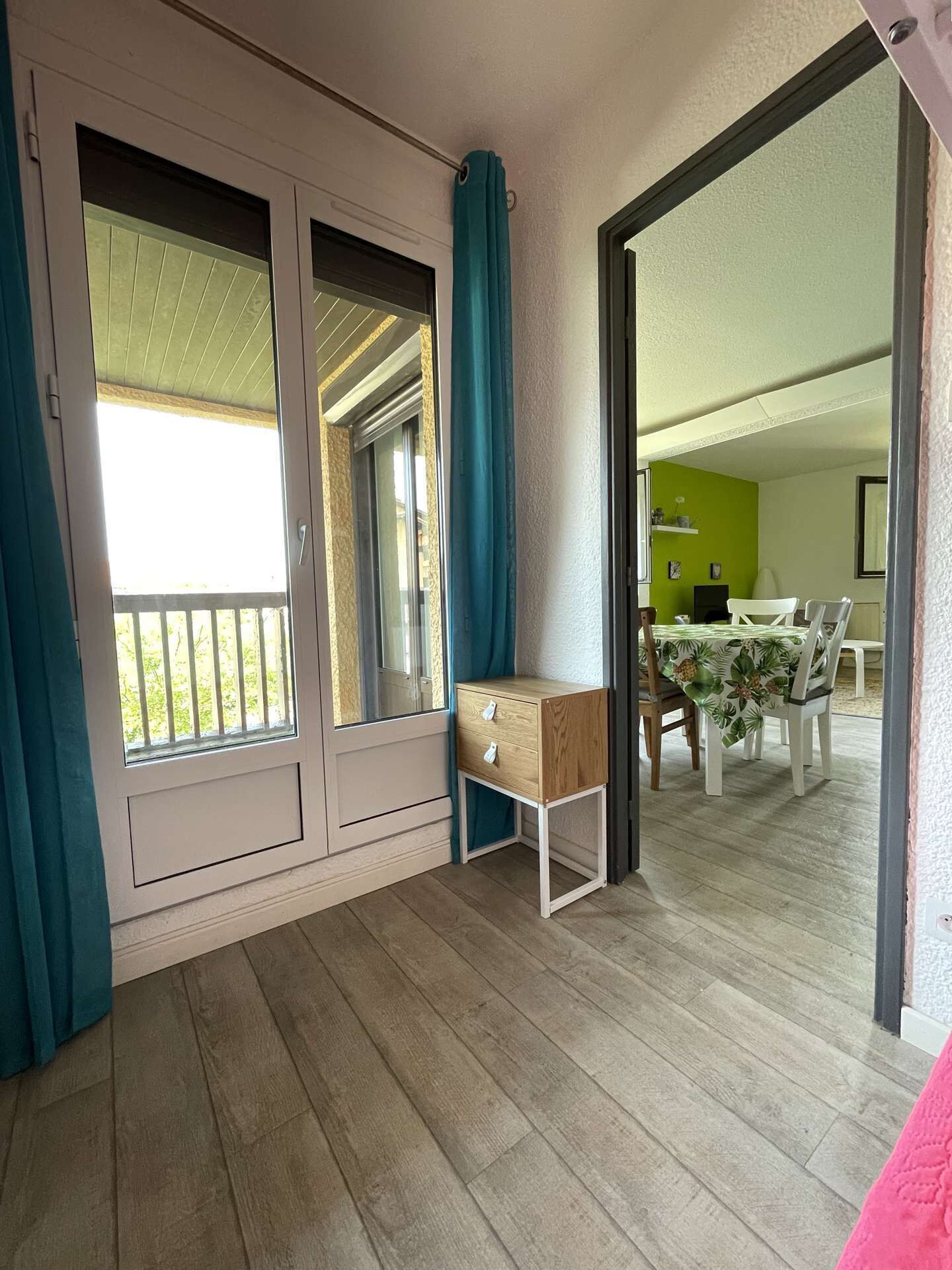 A room with a wooden floor, a small wooden cabinet, a dining table with chairs, and a balcony with a railing.