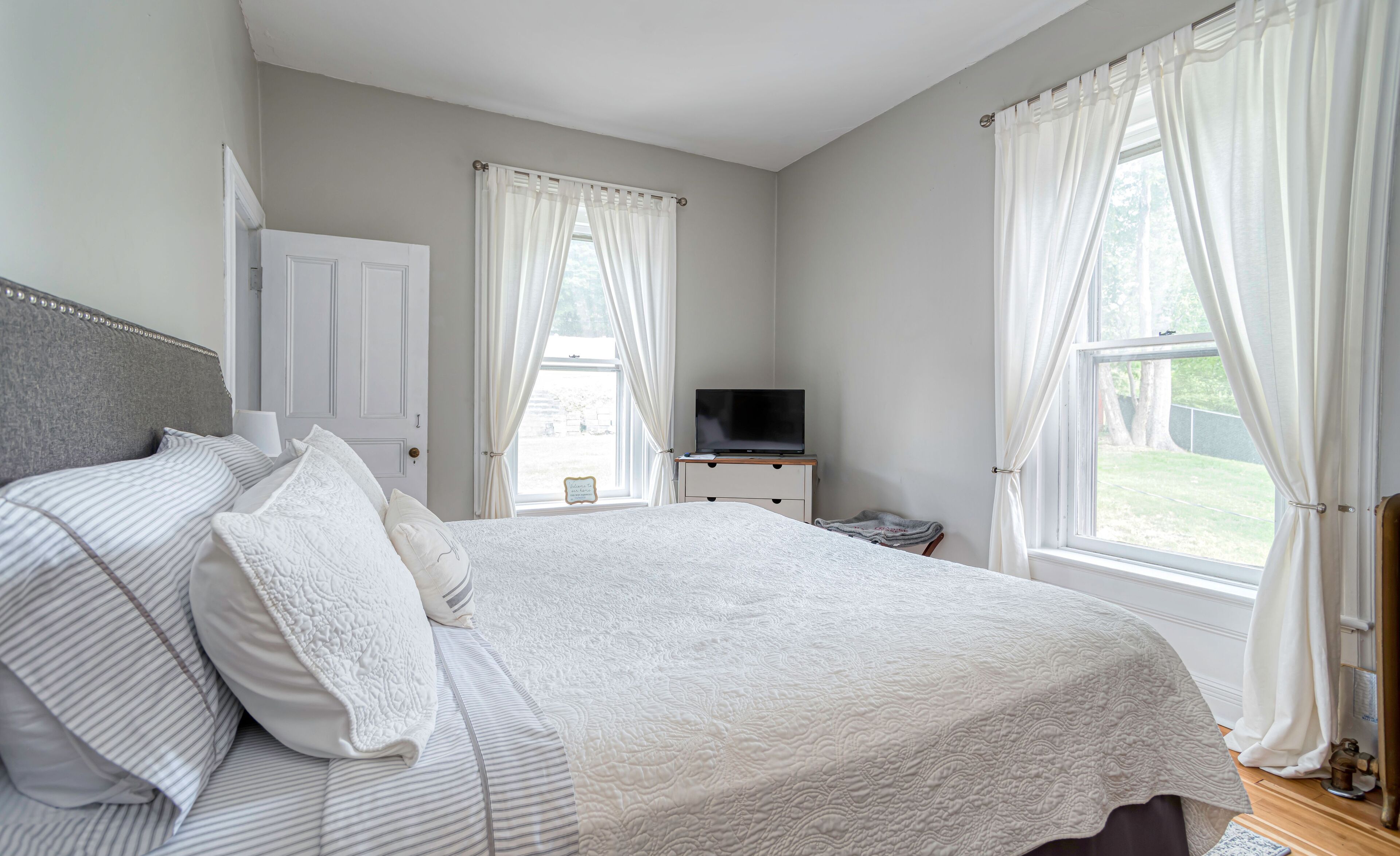 Une chambre à coucher avec un grand lit, un téléviseur posé sur une commode et une fenêtre avec des rideaux.