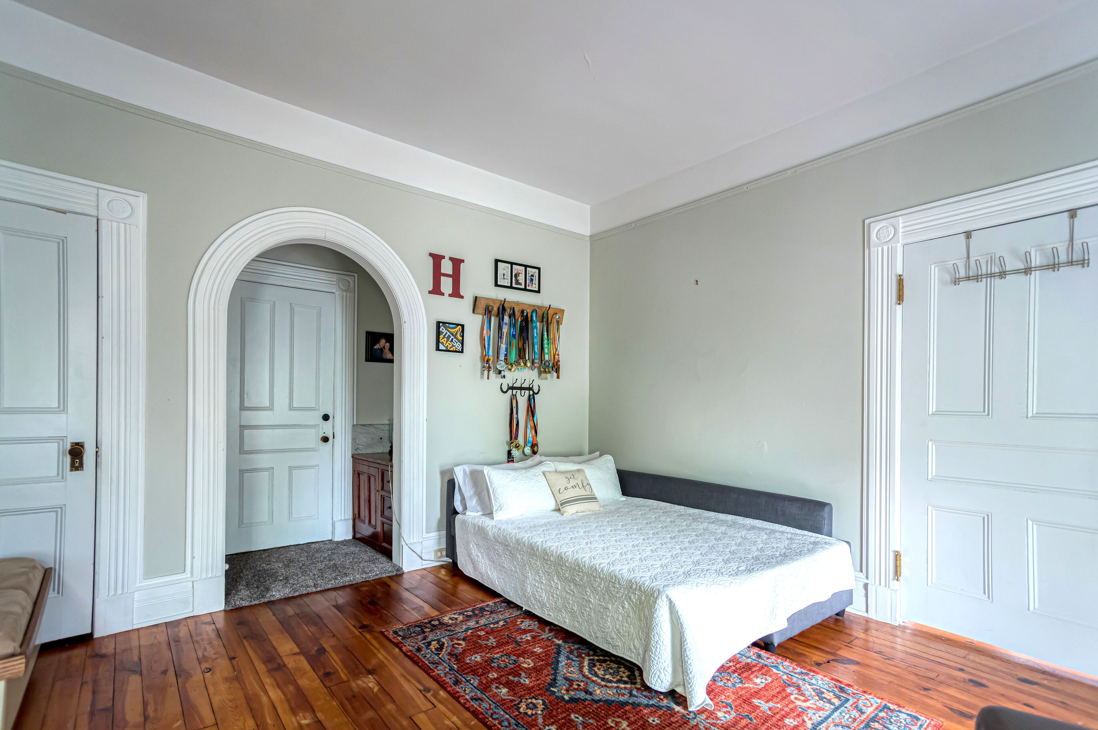 Un dormitorio con una cama, un tapete rojo y azul con estampado, un perchero con bufandas coloridas y una puerta blanca con la letra 'H' en la pared.
