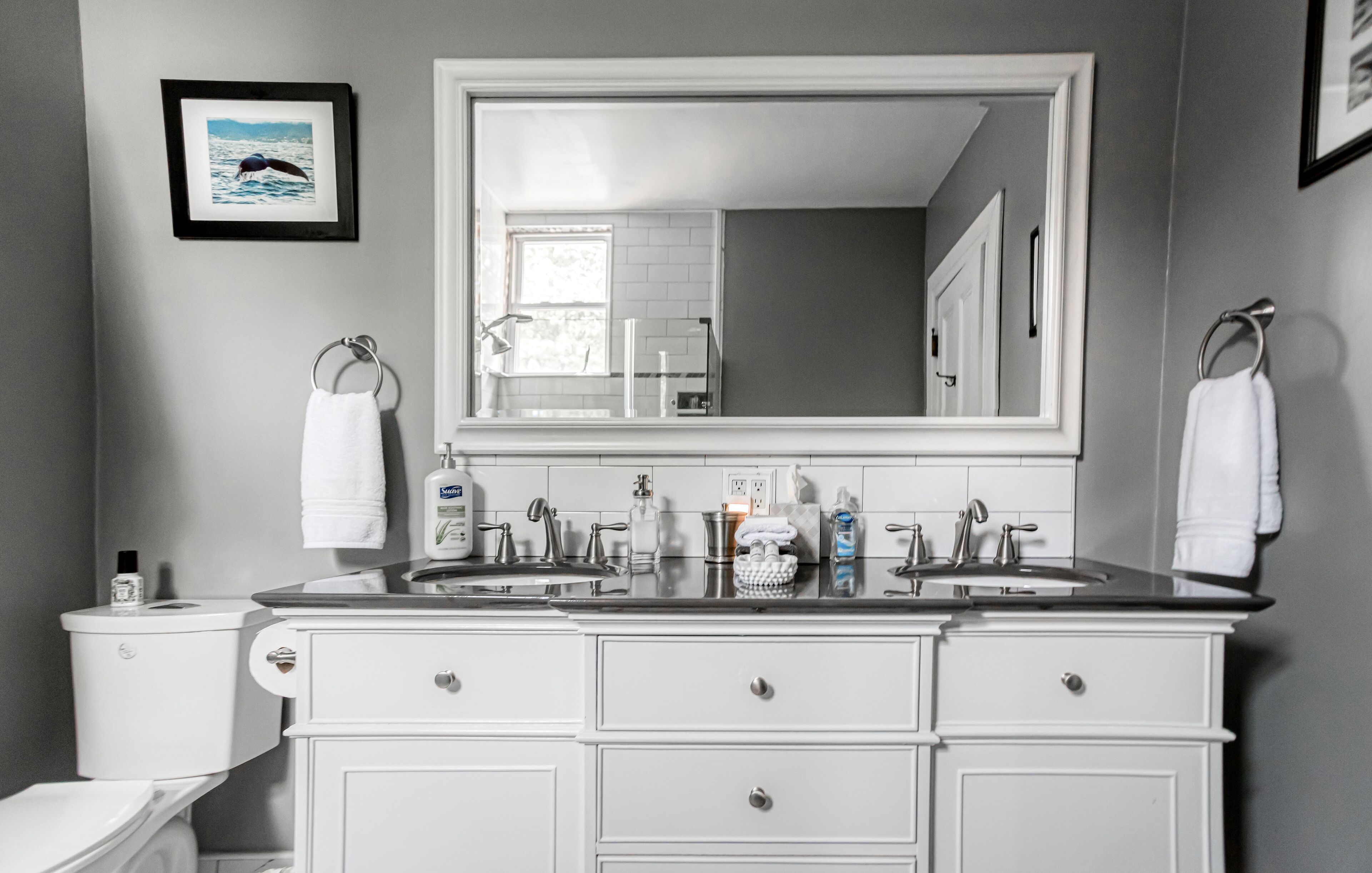 A bathroom with a large mirror, two sinks, white cabinets, and a toilet.