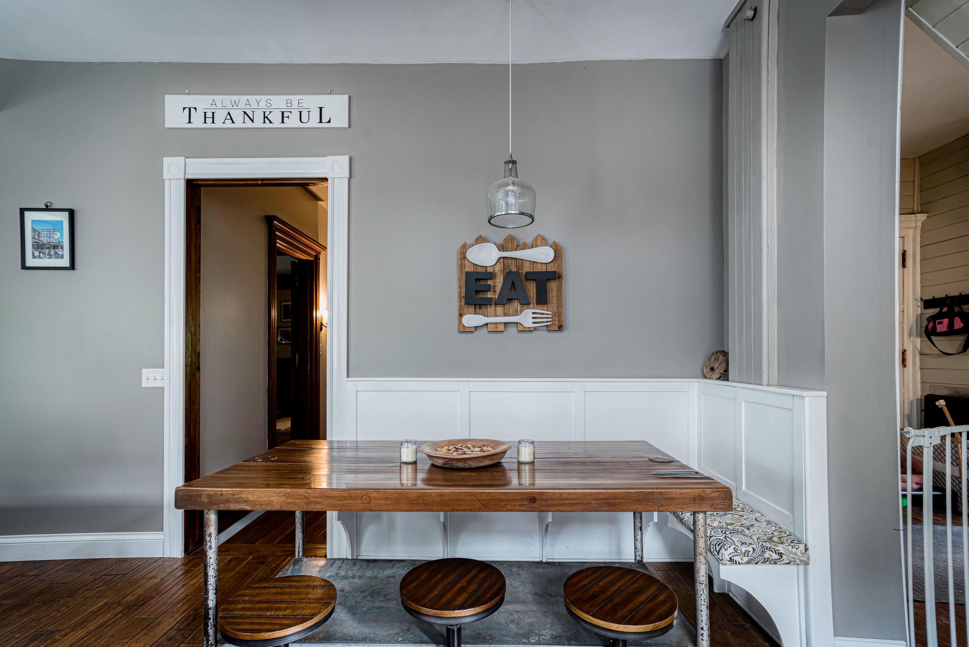 Un comedor con una mesa de madera, tres taburetes, un letrero en la pared que dice "ALWAYS BE THANKFUL" y una lámpara colgante.