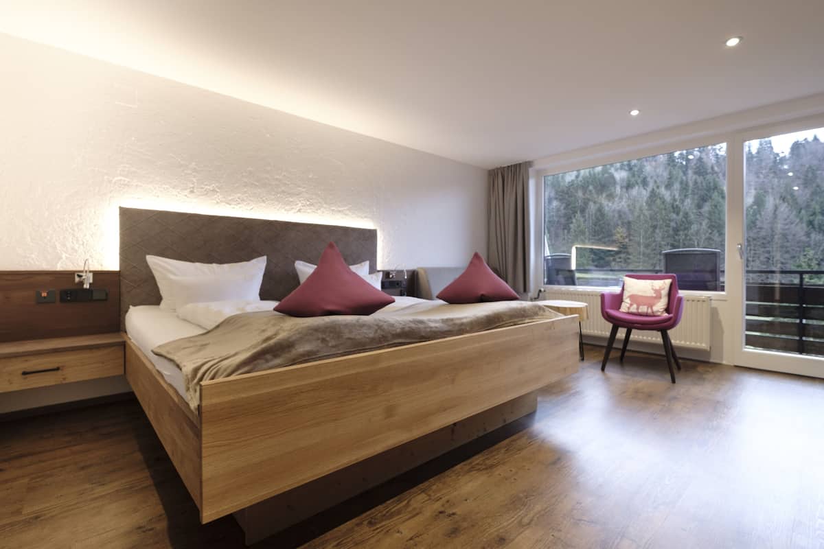 Ein modernes Schlafzimmer mit einem hölzernen Bett, einem Nachttisch, einem Sessel und einem großen Fenster mit Blick auf Bäume.