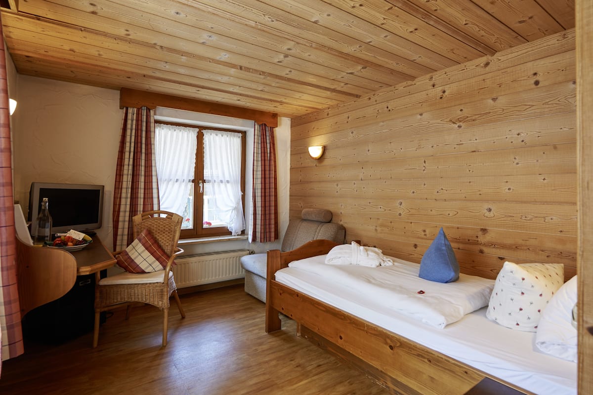 Ein Zimmer mit Holzwänden, einem Bett, einem Sessel, einem Schreibtisch und einem Fenster mit Vorhängen.