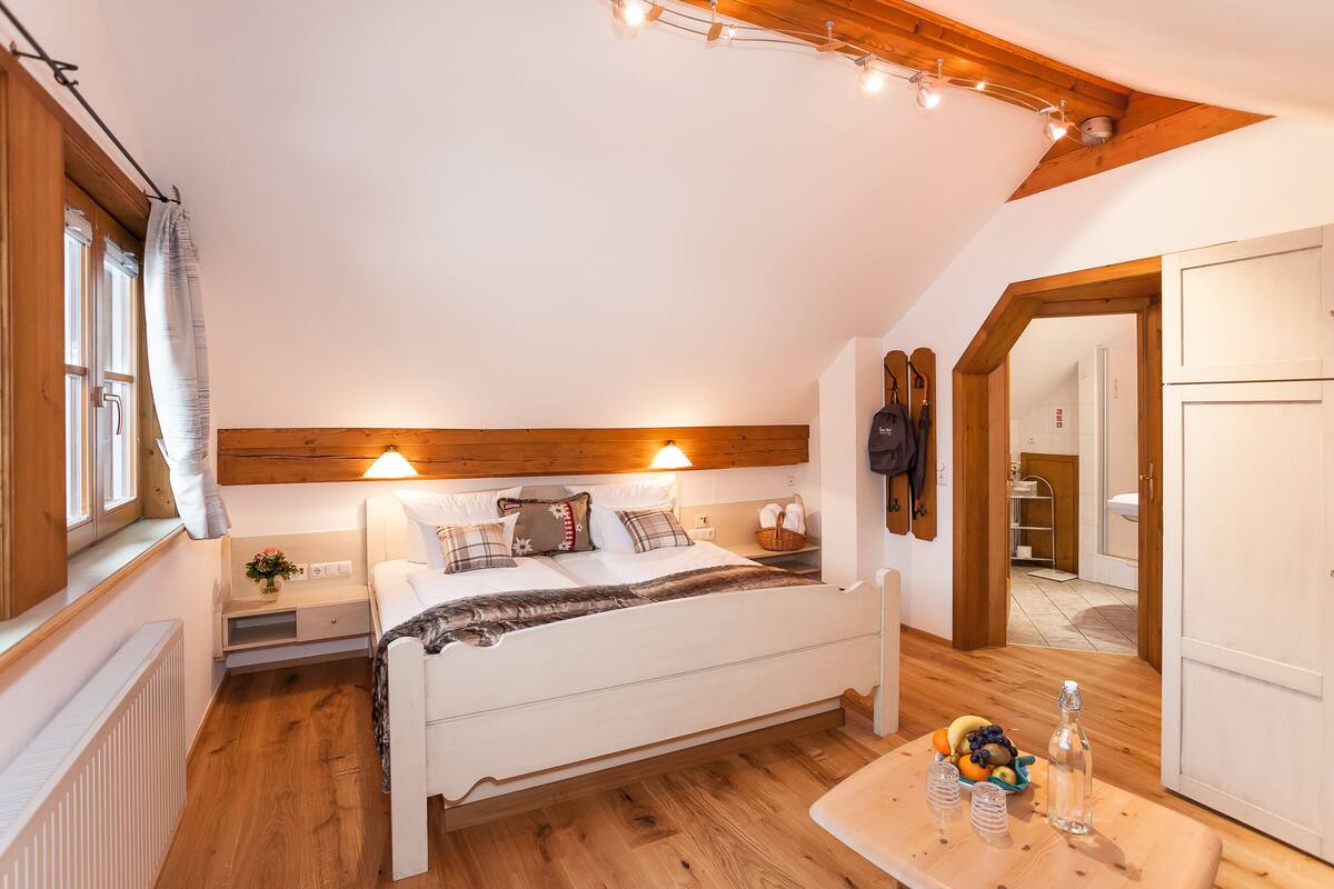 Ein gemütliches Schlafzimmer mit Holzboden, einem Bett mit Kissen, einem Nachttisch und einem Tisch mit Früchten und einer Flasche.