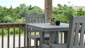 A wooden deck with a table and chairs, surrounded by greenery. - 3-bedroom, 2-bathroom apartment. Walk to beach and access to REAL Watersports (Waves)