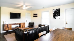 A modern living room with a black leather sofa, wooden dining table, and a mounted flat-screen TV. - 3-bedroom, 2-bathroom apartment. Walk to beach and access to REAL Watersports (Waves)