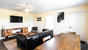 A modern living room with a black leather sofa, a wooden dining table, and a flat-screen TV mounted on the wall.