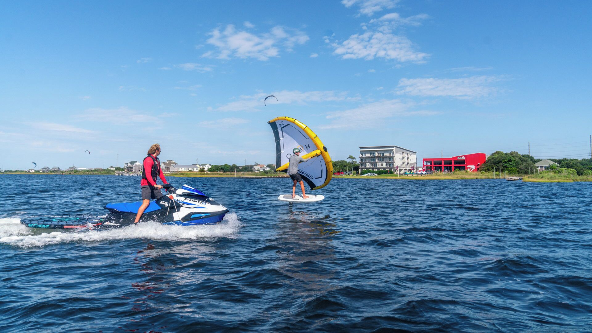 Twee mensen windsurfen op een meer, met gebouwen op de achtergrond.