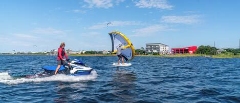 Twee mensen windsurfen op een meer, met gebouwen op de achtergrond.