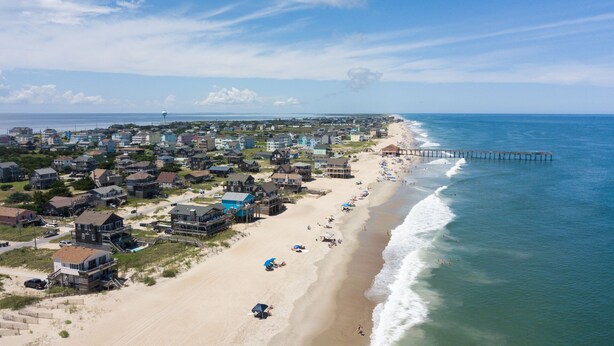 Un pueblo costero con propiedades frente al mar, un muelle que se adentra en el océano y un cielo despejado.