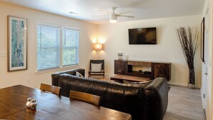 Una sala de estar con un sofá de cuero oscuro, una mesa de comedor de madera y un televisor montado en la pared.