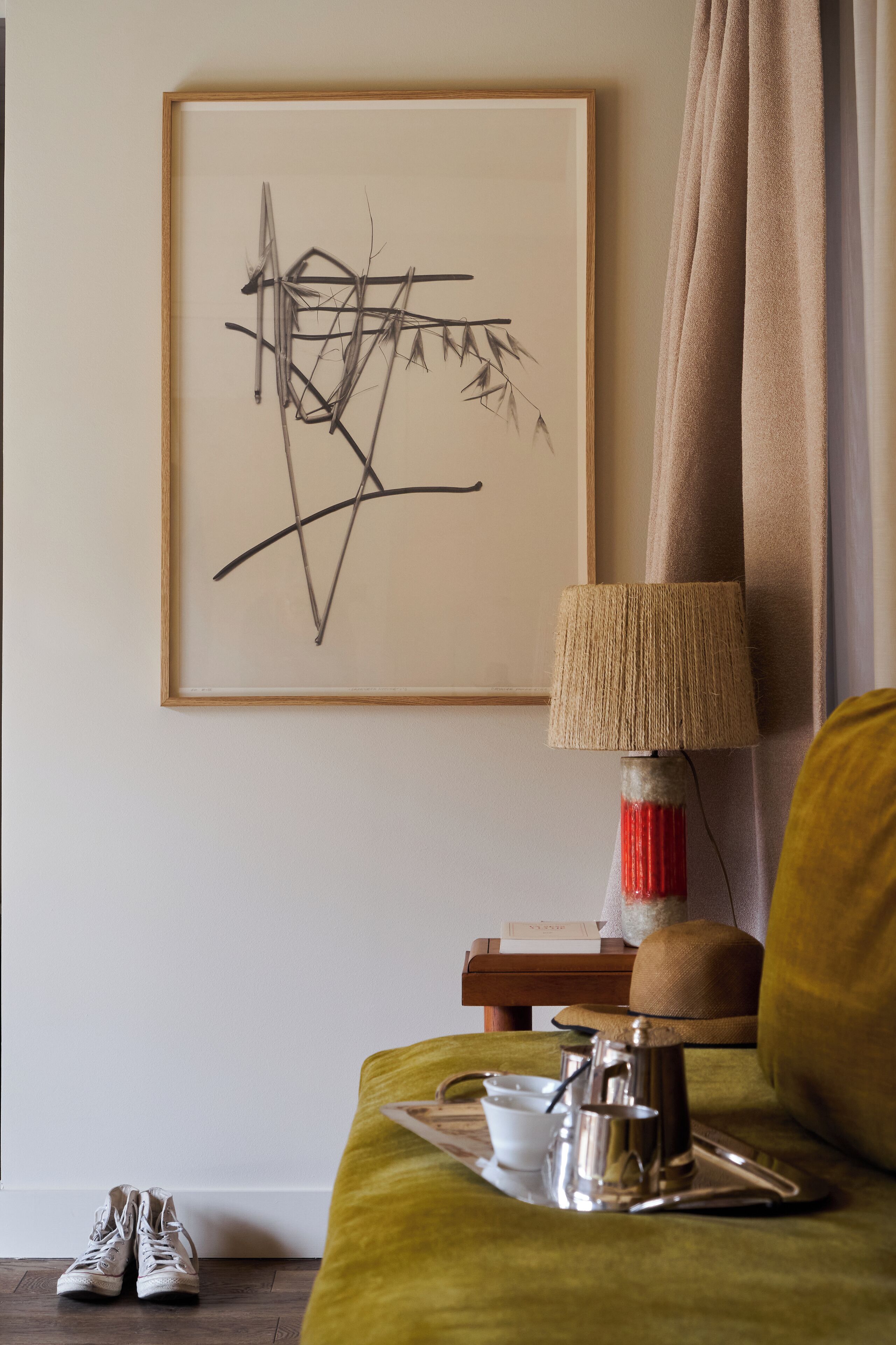 Una habitación con una obra de arte abstracta enmarcada, una lámpara con pantalla texturizada, una bandeja con una taza y una tetera, y un par de zapatos en el suelo.