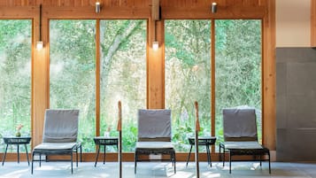 A pool area with wooden ceiling beams, four lounge chairs, and a view of greenery through large windows.
